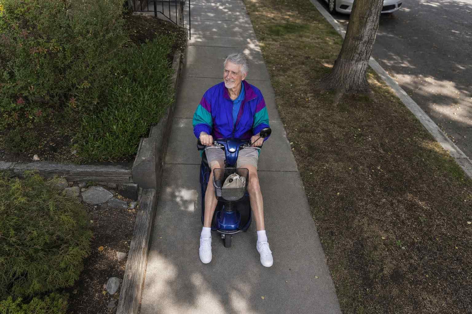 A tall older adult riding a mobility scooter along a sidewalk.