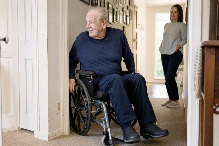 A man in a wheelchair turning through a hallway doorway at home while his adult daughter looks on, the door frame edge visible beside his armrest