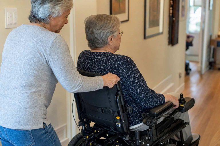 A helper pushing an older adult's electric wheelchair down a hallway.