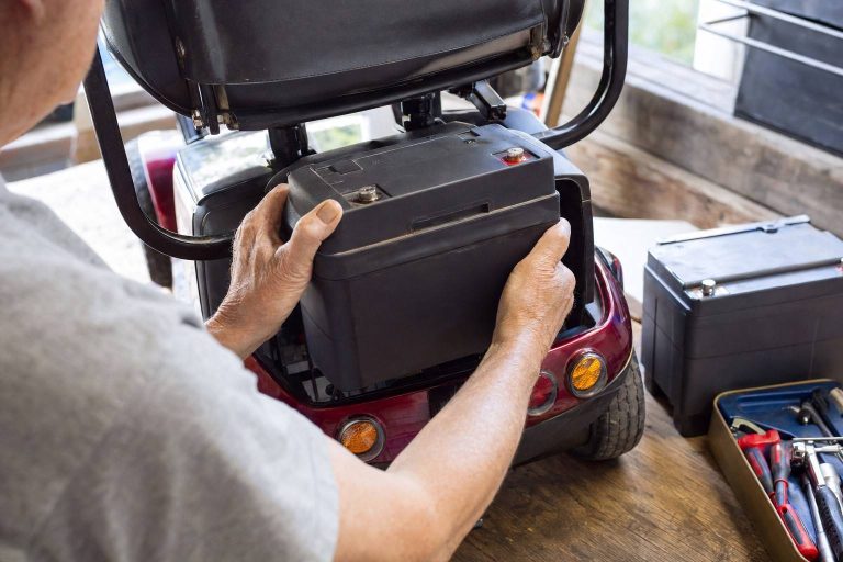 Hands lifting a battery pack from beneath a mobility scooter seat.
