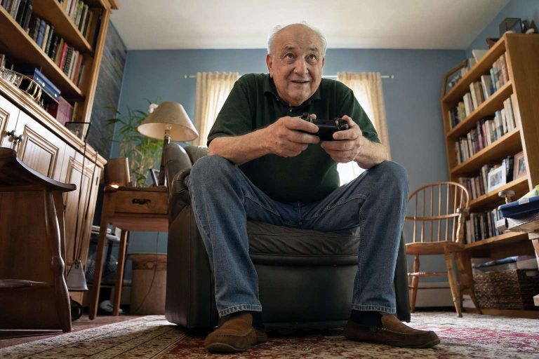An older adult holding a game controller in a den room.