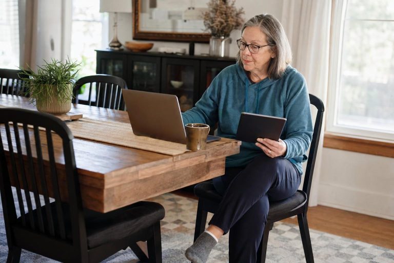 An older adult comparing a tablet and laptop at the dining table.